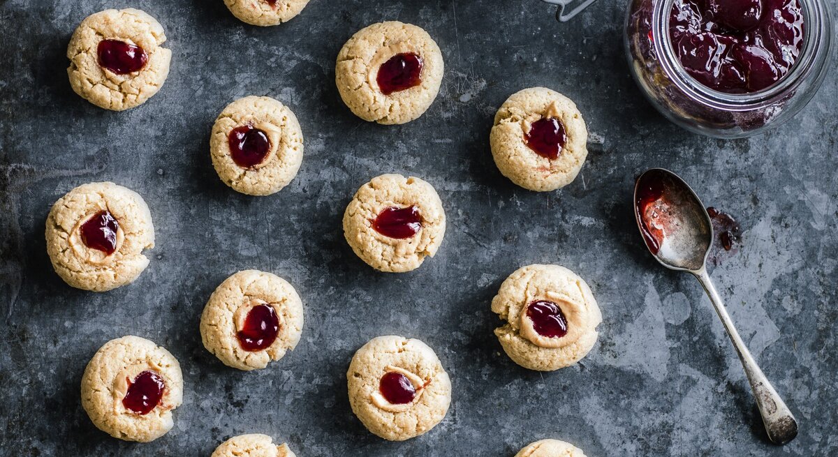 Kekse mit Marmelade und Erdnussbutter auf grauem Hintergrund, rechts ein Glas gefüllt mit Marmelade.