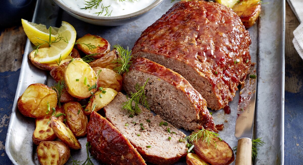 Glasierter Hackbraten mit Kartoffeln auf einem silbernen Blech. Oben auf dem Foto ein Schüsselchen mit einer Soße, rechts unten ein Messer. 