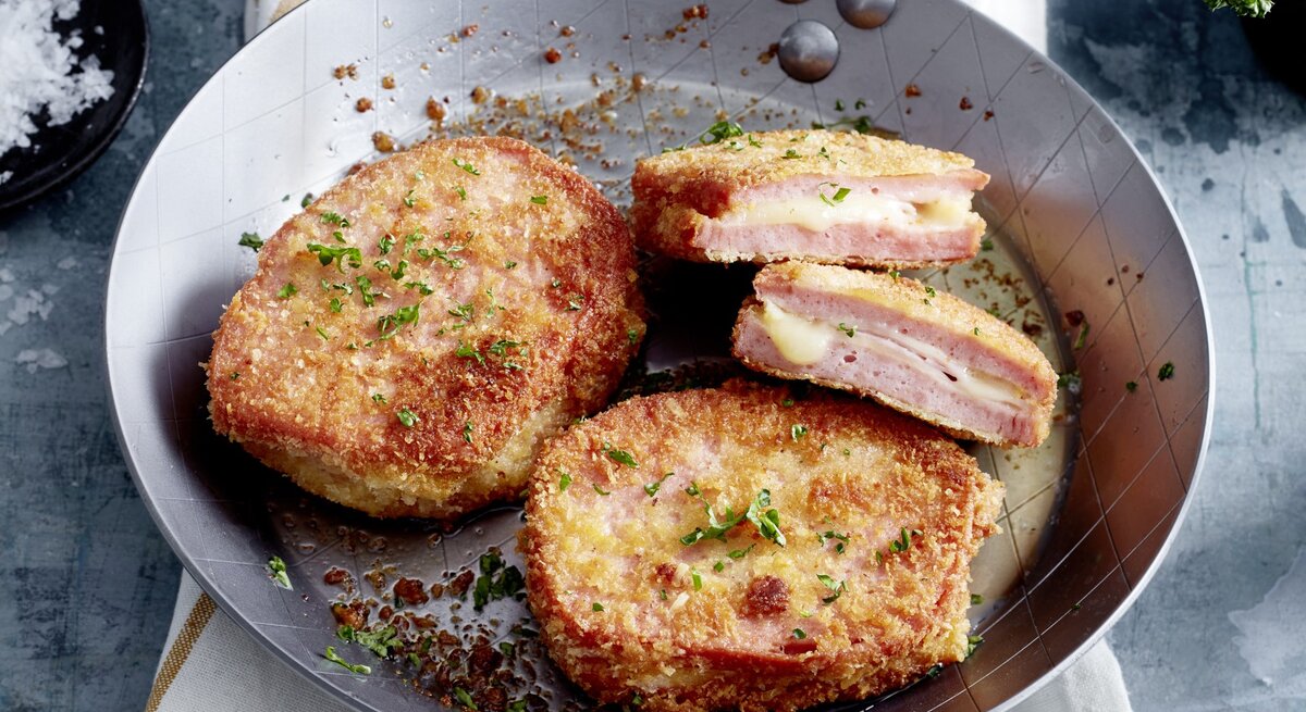 Fleischkäse-Cordon-bleu in einer Pfanne auf einem weißen Tuch auf silbernem Hintergrund.