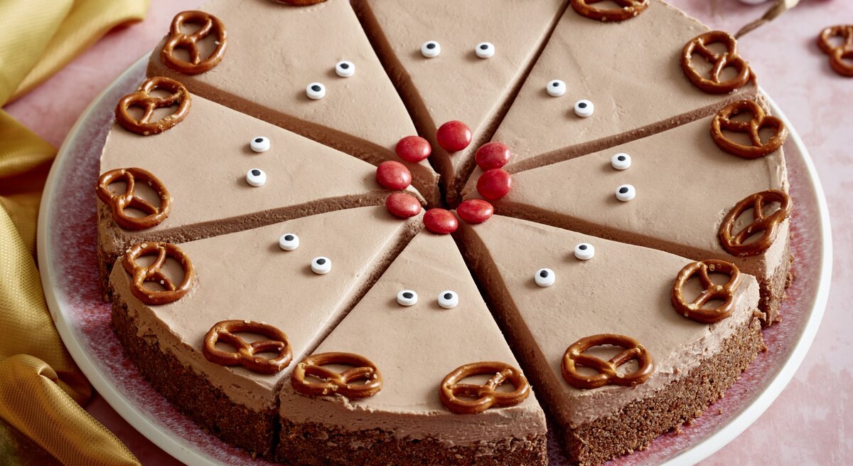 Rudolph-Cheesecake auf einem rosa Teller auf rosa Hintergrund. Links ein gelbes Tuch und rechts Weihnachtskugeln verteilt.