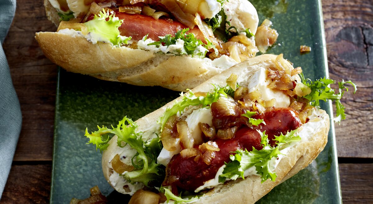 zwei Hotdogs gefüllt mit Camembert und schwarfen Würsten auf grünem Teller.