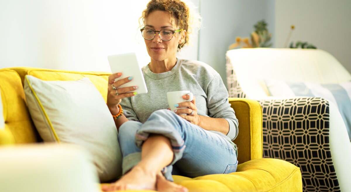 Eine Person mit lockigen Haaren sitzt entspannt auf einem gelben Sofa mit Kissen, hält ein Tablet und eine Tasse in einem hellen Raum.