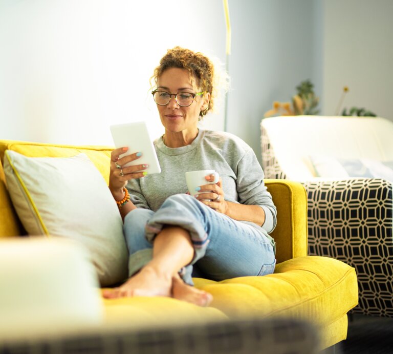 Eine Person mit lockigen Haaren sitzt entspannt auf einem gelben Sofa mit Kissen, hält ein Tablet und eine Tasse in einem hellen Raum. | © Adobe Stock/ simona