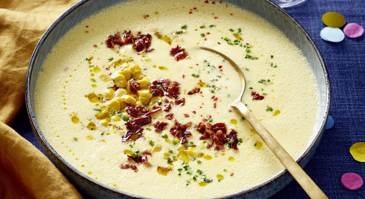 Mais-Cremesuppe in einer grauen Schüssel auf dunkelblauem Hintergrund. In der Schüssel ein goldener Löffel und links davon ein gelbes Geschirrtuch.