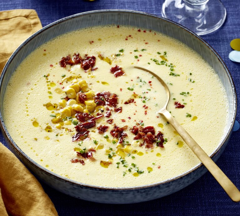 Mais-Cremesuppe in einer grauen Schüssel auf dunkelblauem Hintergrund. In der Schüssel ein goldener Löffel und links davon ein gelbes Geschirrtuch. | © EAT CLUB/Isabell Triemer