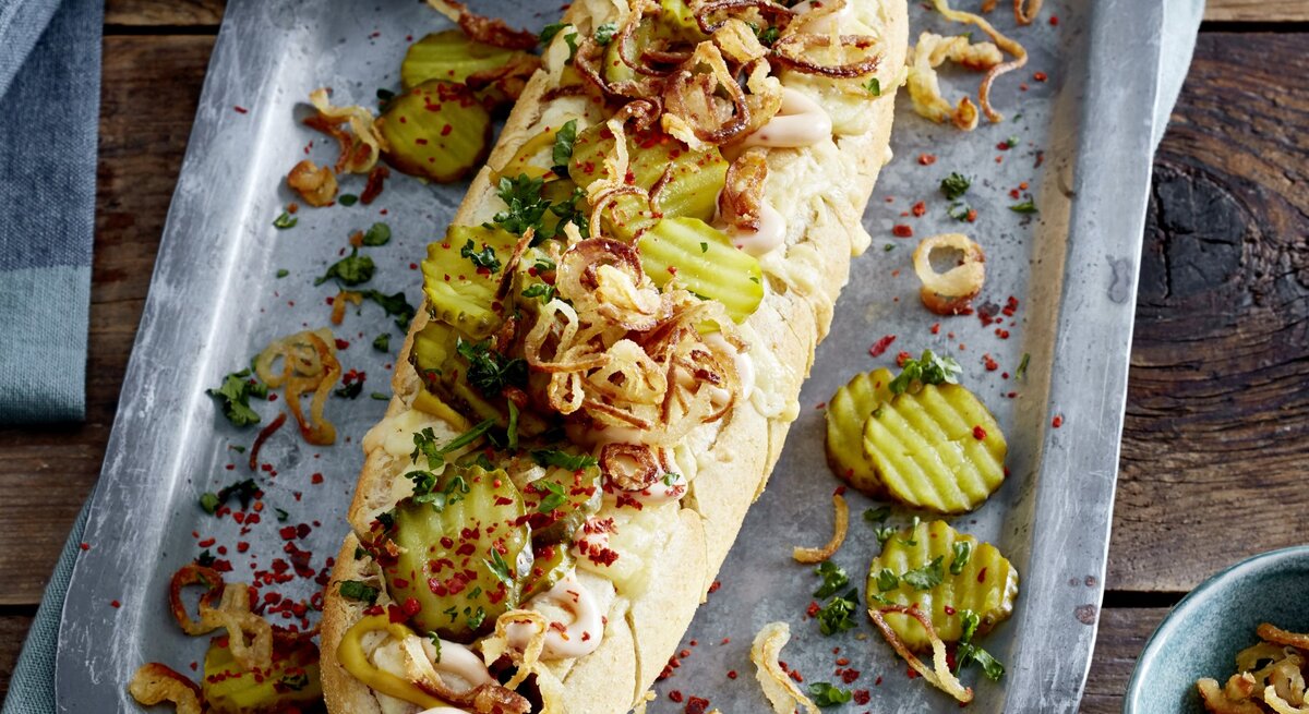 Hotdog ofengebacken im Kräuterbaguette auf einem silberfarbenen Blech auf hölzernem Hintergrund. 