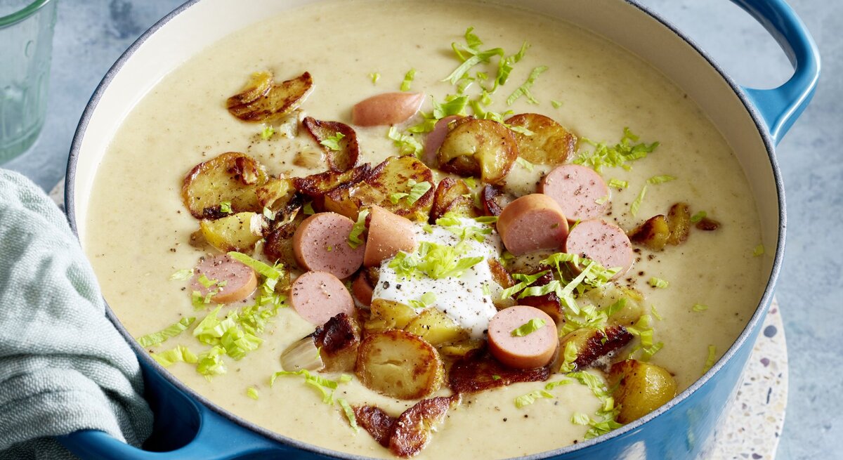 Bratkartoffel-Suppe mit Würstchen in einem blauen Topf auf grauem Hintergrund. 