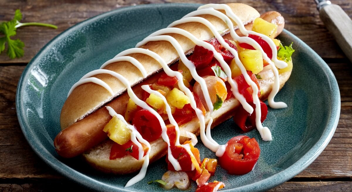 Hotdog mit Ananas & Jalapeños auf einem dunkelblauen Teller auf hölzernem Hintergrund. Rechts von dem Teller eine Gabel. 