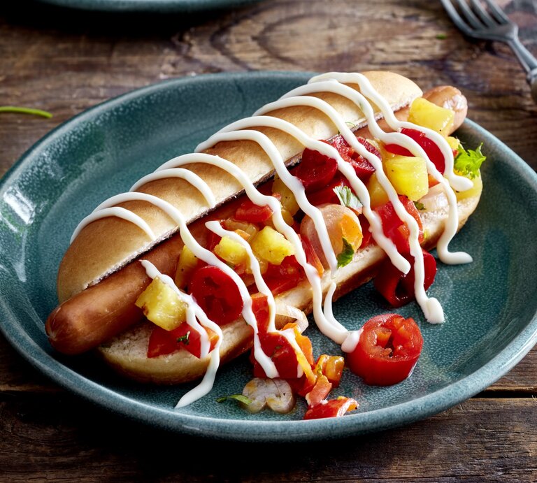 Hotdog mit Ananas & Jalapeños auf einem dunkelblauen Teller auf hölzernem Hintergrund. Rechts von dem Teller eine Gabel.  | © EAT CLUB/Isabell Triemer