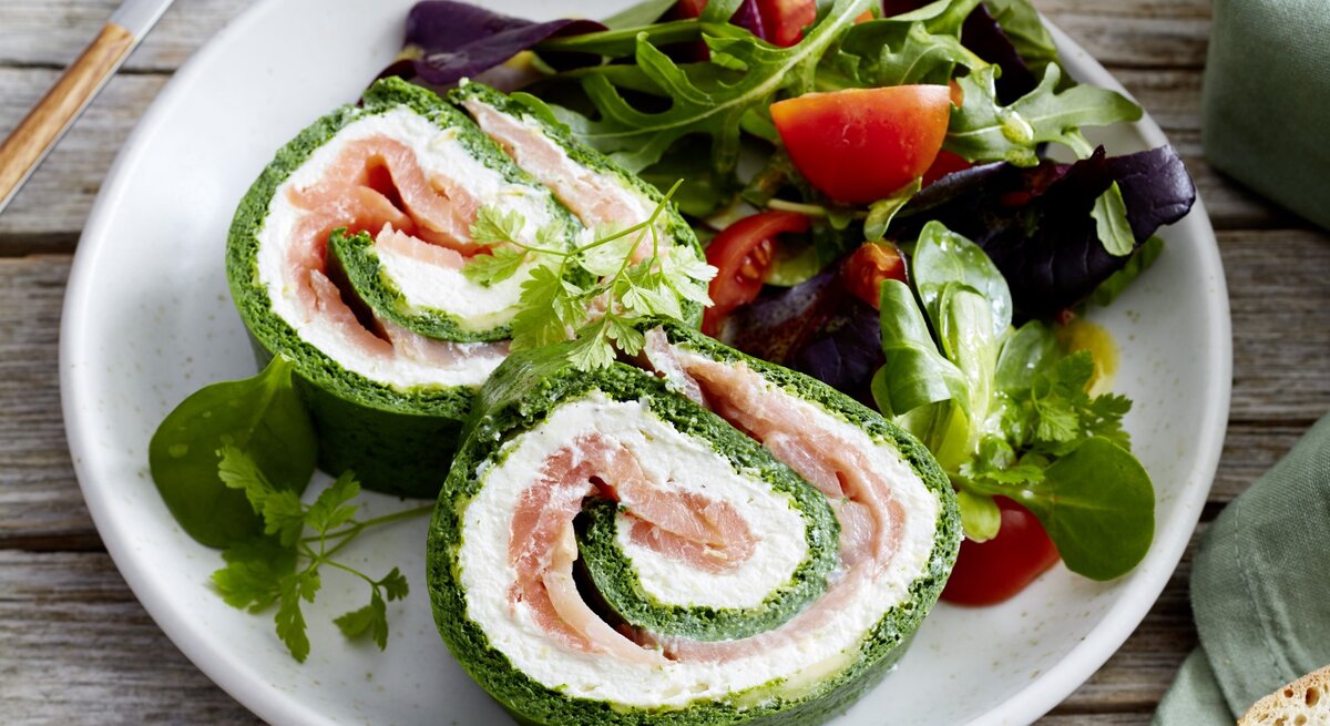 Spinat-Lachs-Roulade auf einem weißen Teller angerichtet  mit Salat auf hölzernem Hintergrund. Links davon Besteck, rechts Brot.