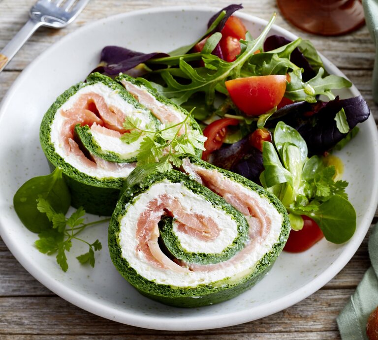 Spinat-Lachs-Roulade auf einem weißen Teller angerichtet  mit Salat auf hölzernem Hintergrund. Links davon Besteck, rechts Brot. | © EAT CLUB/Isabell Triemer