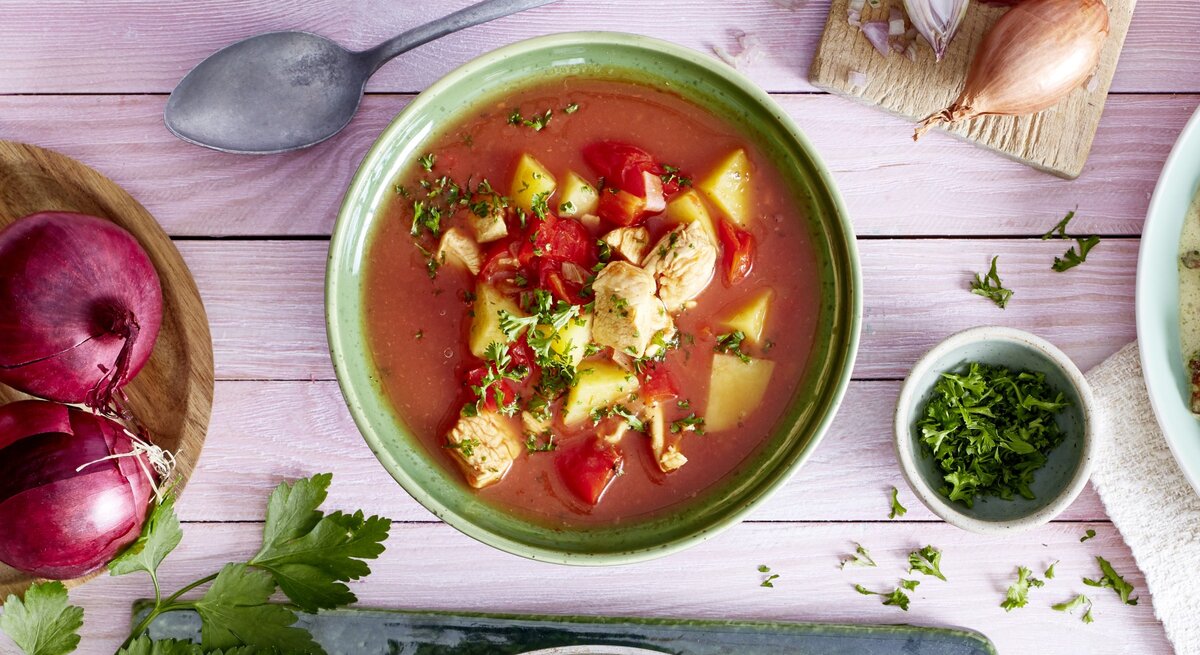 Tomatensuppe mit Huhn in einer grünen Schüssel auf rosa Hintergrund. Links davon Zwiebeln, rechts Schalotten und Petersilie. 