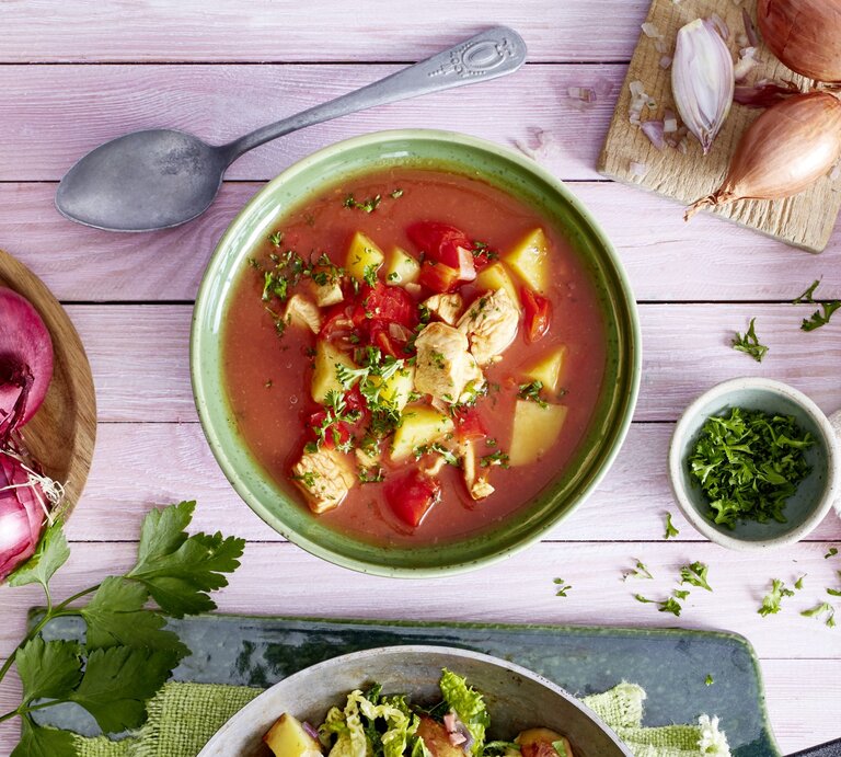 Tomatensuppe mit Huhn in einer grünen Schüssel auf rosa Hintergrund. Links davon Zwiebeln, rechts Schalotten und Petersilie.  | © EAT CLUB / Isabell Triemer