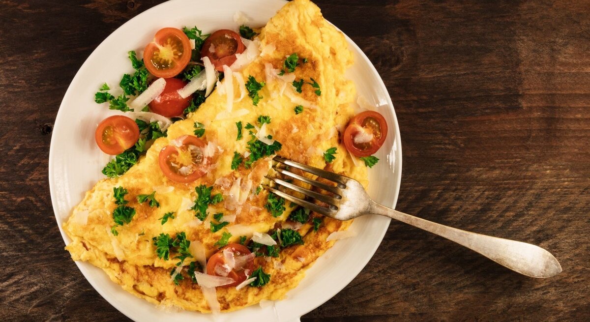 Ein goldbraunes Omelett mit Kirschtomaten, Petersilie und Parmesan auf einem weißen Teller mit einer Gabel auf dunklem Holztisch.