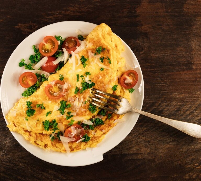 Ein goldbraunes Omelett mit Kirschtomaten, Petersilie und Parmesan auf einem weißen Teller mit einer Gabel auf dunklem Holztisch. | © Shutterstock/lateresca