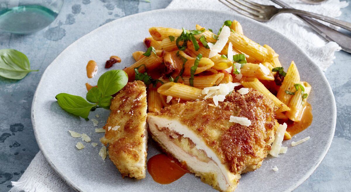 Italienisches Hähnchen-Cordon-bleu auf einem grauen Teller auf einem grauen Geschirrtuch auf grauem Hintergrund. Rechts oben drei Gabeln, links oben Wasser und Basilikum verteilt.
