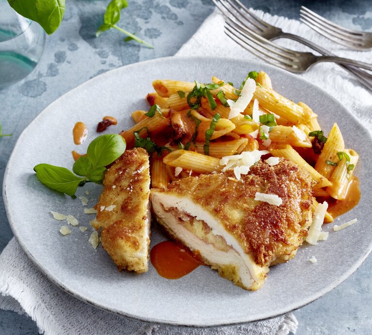 Italienisches Hähnchen-Cordon-bleu auf einem grauen Teller auf einem grauen Geschirrtuch auf grauem Hintergrund. Rechts oben drei Gabeln, links oben Wasser und Basilikum verteilt. | © EAT CLUB/Isabell Triemer