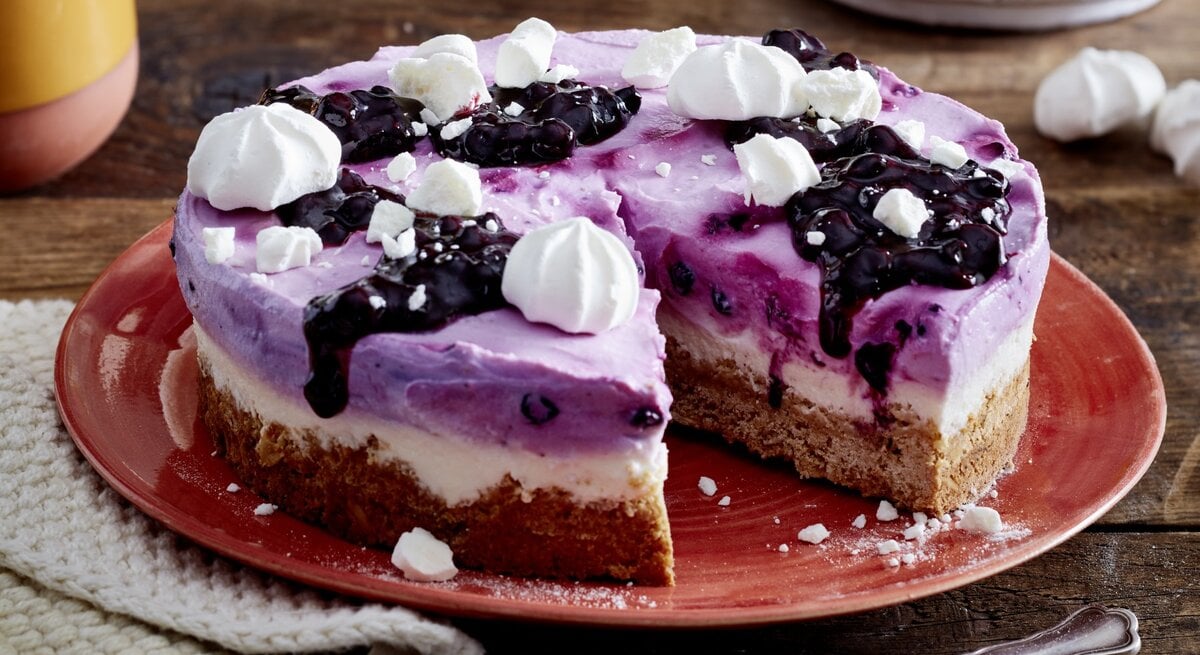 Heidelbeer-Skyr-Torte auf einem roten Teller auf hölzernem Hintergrund. Links auf dem Foto ein weißes Geschirrtuch und unten auf dem Foto Dessertgabeln. 