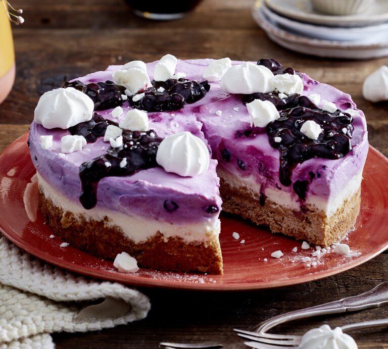 Heidelbeer-Skyr-Torte auf einem roten Teller auf hölzernem Hintergrund. Links auf dem Foto ein weißes Geschirrtuch und unten auf dem Foto Dessertgabeln.  | © EAT CLUB / Isabell Triemer