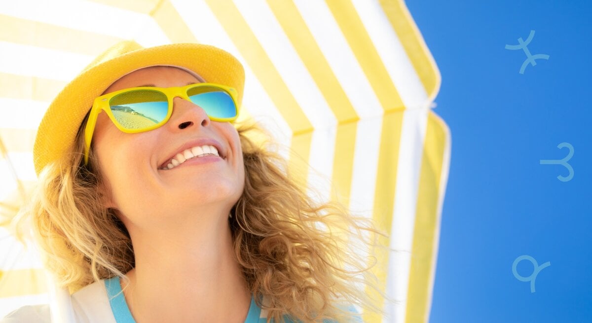 Das Bild zeigt eine lächelnde Frau mit lockigen blonden Haaren, die eine gelbe Sonnenhut und gelbe Sonnenbrille trägt. Sie steht unter einem gelb-weiß gestreiften Sonnenschirm vor einem klaren, blauen Himmel. Auf dem Himmel sind leicht transparente Symbole verschiedener Sternzeichen zu sehen. Die Stimmung ist sommerlich und fröhlich.