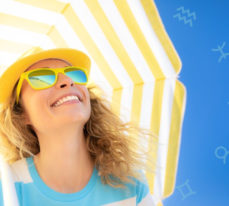 Das Bild zeigt eine lächelnde Frau mit lockigen blonden Haaren, die eine gelbe Sonnenhut und gelbe Sonnenbrille trägt. Sie steht unter einem gelb-weiß gestreiften Sonnenschirm vor einem klaren, blauen Himmel. Auf dem Himmel sind leicht transparente Symbole verschiedener Sternzeichen zu sehen. Die Stimmung ist sommerlich und fröhlich. | © Adobe Stock/ Sunny studio und Chikovnaya via Canva.com [M]