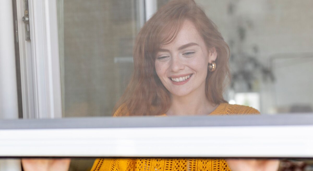 Eine junge Frau mit roten Haaren und goldenen Ohrringen lächelt beim Fensteröffnen in einem hellen, gemütlichen Raum.
