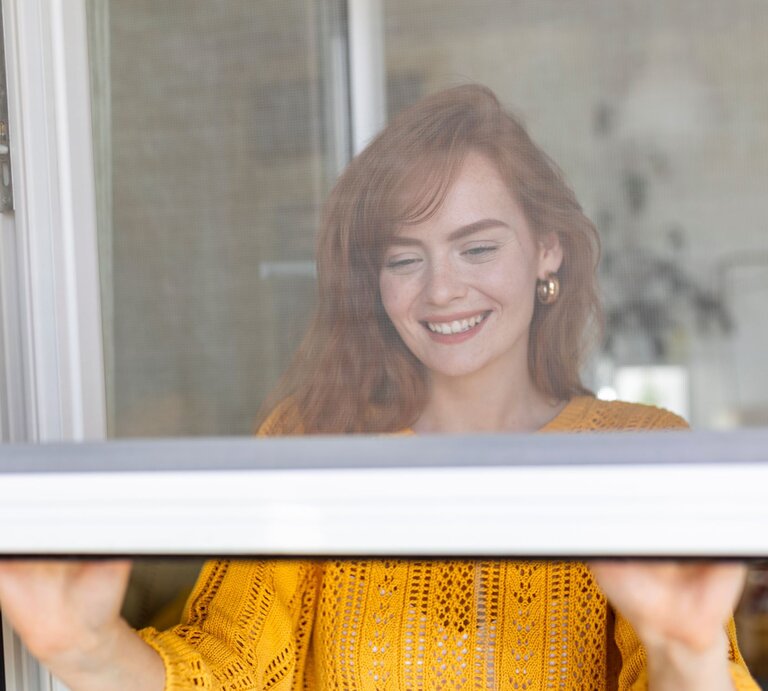 Eine junge Frau mit roten Haaren und goldenen Ohrringen lächelt beim Fensteröffnen in einem hellen, gemütlichen Raum. | © Adobe Stock/ Graphicroyalty