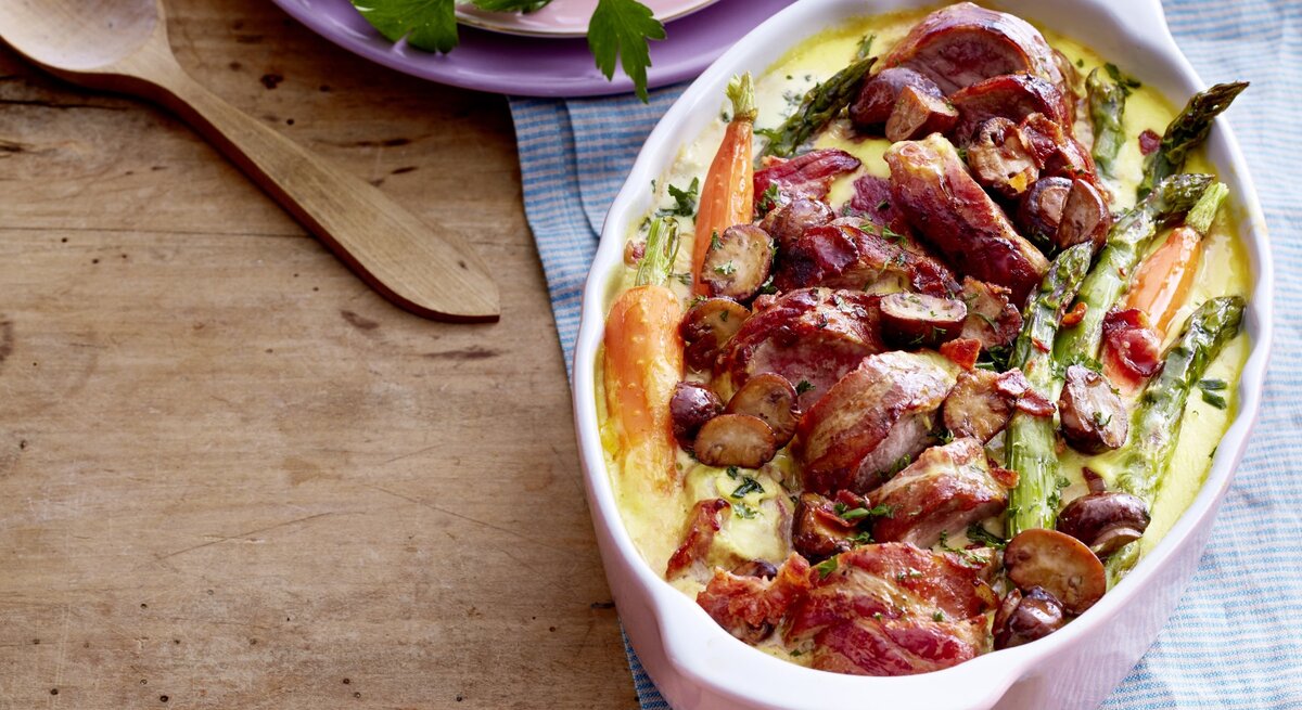 Schweinefilet mit Frühlingsgemüse in einer weißen Auflaufform, daneben Teller mit Petersilie und Holzlöffel auf Holzuntergrund. 