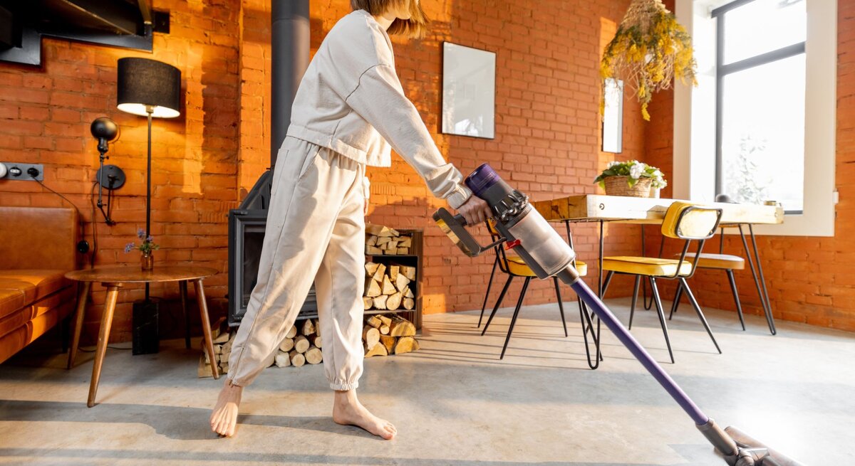  Das Bild zeigt eine Person in einem modern gestalteten Wohnraum mit Ziegelmauerwerk, die einen kabellosen Dyson-Staubsauger benutzt. Die Person ist barfuß und trägt lässige, bequeme Kleidung. Sie scheint den Staubsauger zu führen, um den hellgrauen Betonboden zu reinigen. Im Hintergrund ist ein gemütlich eingerichteter Raum mit einem braunen Ledersofa, einem runden Holztisch und einer Sitzgruppe mit gelben Stühlen an einem hölzernen Tisch zu sehen. Über dem Tisch hängt eine Pflanze von der Decke, und ein Fenster lässt natürliches Licht herein. Die warmen Farben und das Holzfeuer im Kamin verleihen dem Raum eine einladende und wohnliche Atmosphäre.
