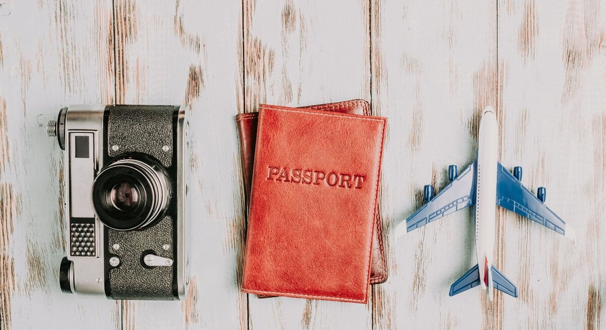 Es sind eine rote Geldbörse, ein Fotoapparat und ein Spielzeugflugzeug liegend auf weiß-grauem Untergrund zu sehen.