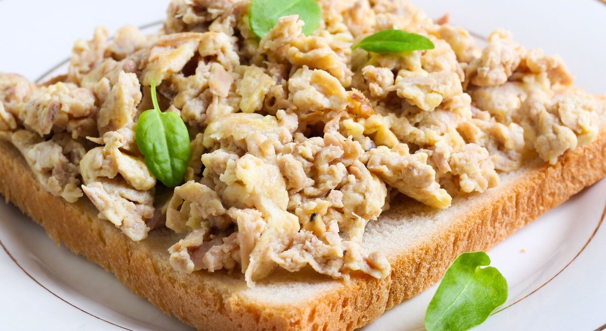 Ein belegtes Toastbrot mit Thunfisch-Rührei garniert mit frischen grünen Blättern, wahrscheinlich Feldsalat, auf einem weißen Teller mit einem goldenen Rand, serviert auf einer gelb gemusterten Tischdecke.