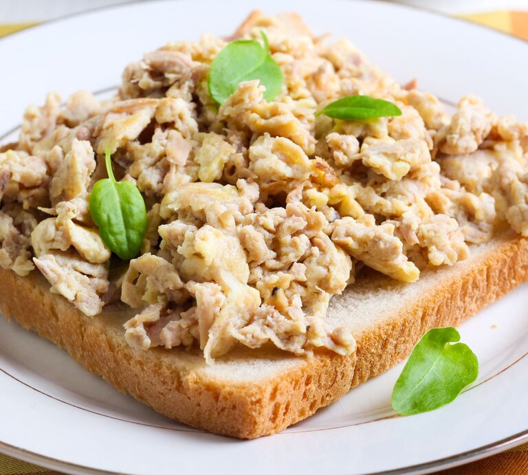 Ein belegtes Toastbrot mit Thunfisch-Rührei garniert mit frischen grünen Blättern, wahrscheinlich Feldsalat, auf einem weißen Teller mit einem goldenen Rand, serviert auf einer gelb gemusterten Tischdecke. | © AdobeStock/manyakotic