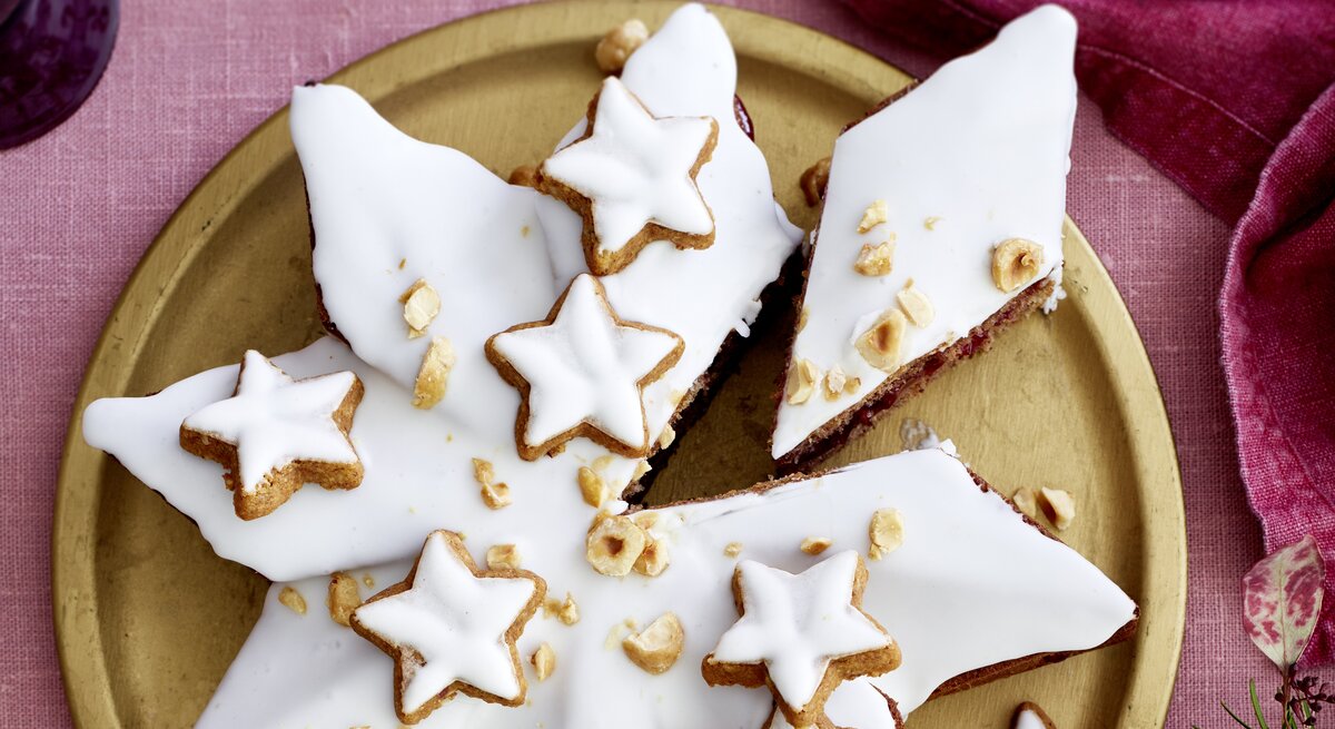 Weihnachtlicher Zimtsternkuchen mit glänzendem Zuckerguss, Zimtsternen und Haselnüssen auf edler Goldplatte, vor rosa Tischwäsche und Deko.