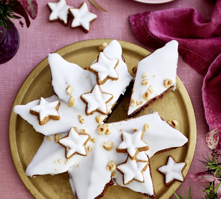 Weihnachtlicher Zimtsternkuchen mit glänzendem Zuckerguss, Zimtsternen und Haselnüssen auf edler Goldplatte, vor rosa Tischwäsche und Deko. | © EAT CLUB / Isabell Triemer