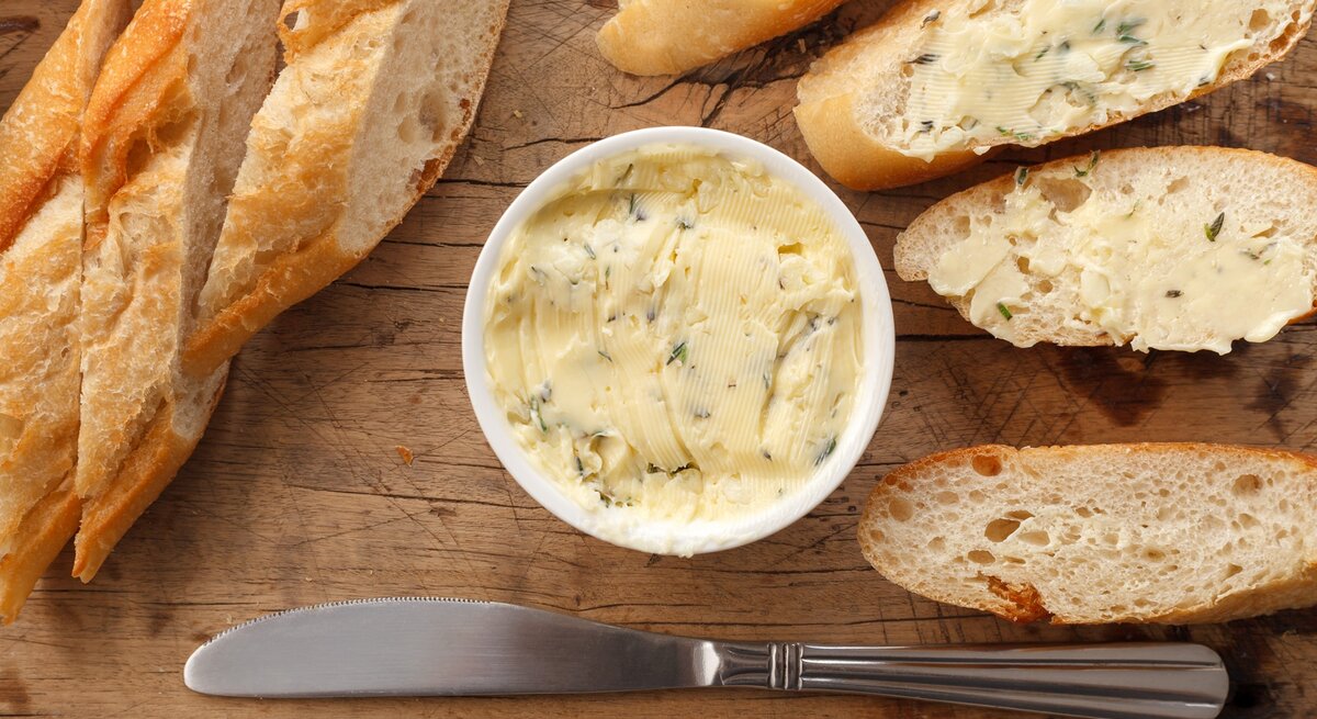 Butter mit Zitronenverbene in einer kleinen runden Form / Baguette ganz und aufgeschnitten / Streichmesser aus Metall / Untergrund grobes Holz