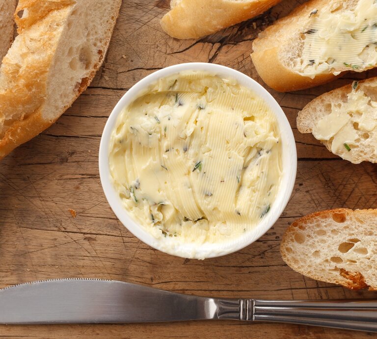 Butter mit Zitronenverbene in einer kleinen runden Form / Baguette ganz und aufgeschnitten / Streichmesser aus Metall / Untergrund grobes Holz | © Shutterstock / rukxstockphoto