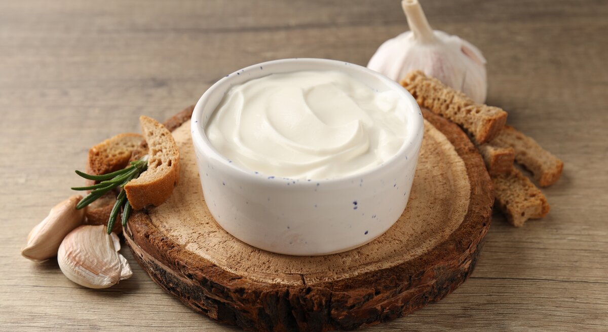 Weiße Schale gefüllt mit Knoblauchsoße auf Holzbrett. Daneben liegen einige Knoblauchzehen, eine Knoblauchknolle und brot-Sticks. 