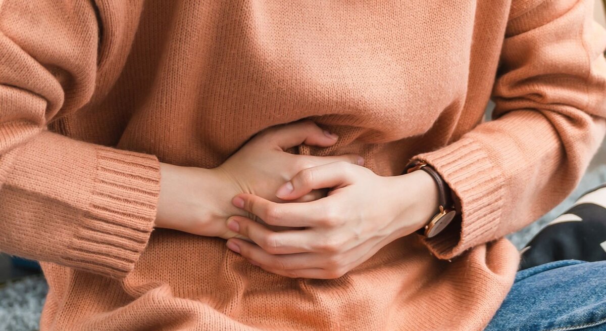  Das Bild zeigt den Mittelteil einer Person, die einen pfirsichfarbenen Strickpullover und eine Uhr trägt und sich mit gefalteten Händen auf dem Bauch hält, was möglicherweise auf Bauchschmerzen oder Unbehagen hinweist.