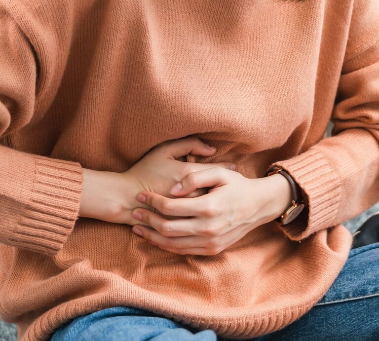  Das Bild zeigt den Mittelteil einer Person, die einen pfirsichfarbenen Strickpullover und eine Uhr trägt und sich mit gefalteten Händen auf dem Bauch hält, was möglicherweise auf Bauchschmerzen oder Unbehagen hinweist. | © iStock.com/Sitthiphong