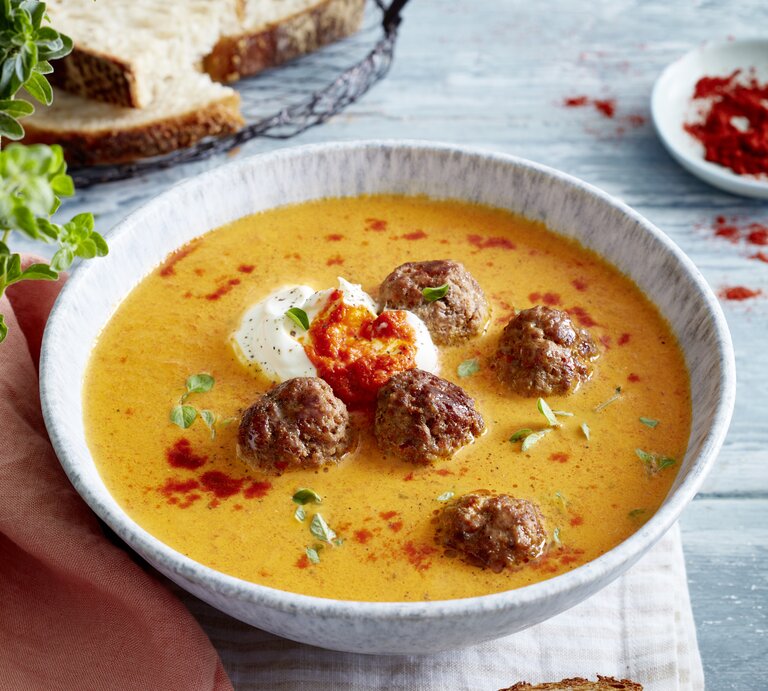 Weiße Schüssel mit cremiger Paprikasuppe mit Hackbällchen und einem Klecks Schmand auf einem gedeckten Esstisch. | © EAT CLUB/Isabell Triemer