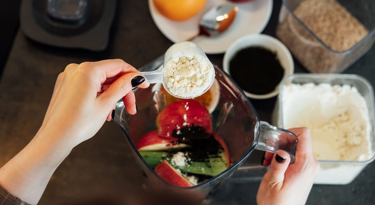 Zwei Hände füllen gesundes Obst und Gemüse sowie Kollagenpulver in einen Mixer.