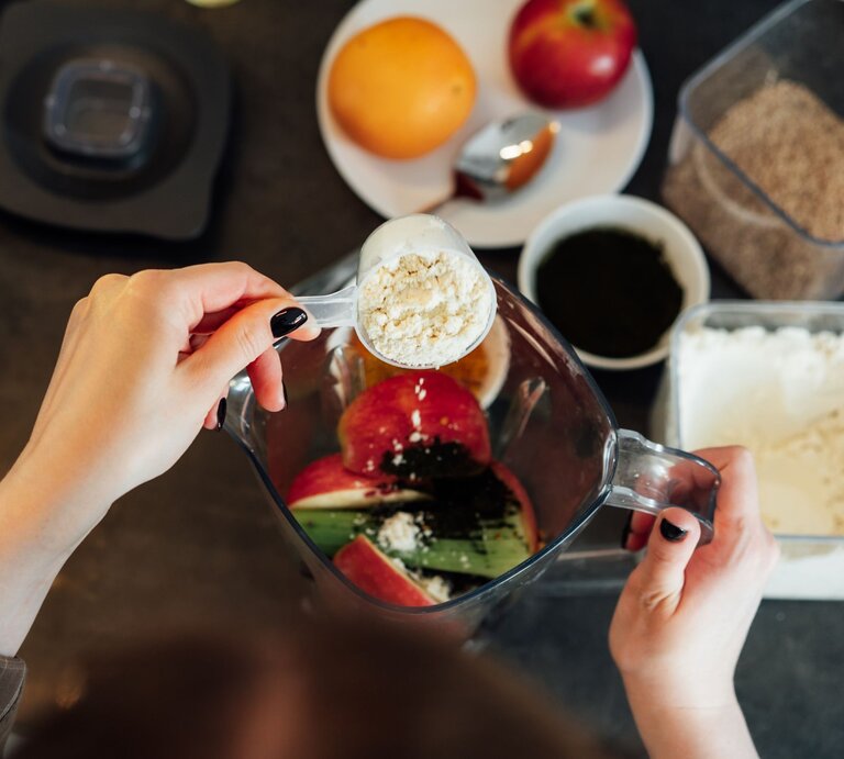 Zwei Hände füllen gesundes Obst und Gemüse sowie Kollagenpulver in einen Mixer. | © gettyimages/ OKrasyuk