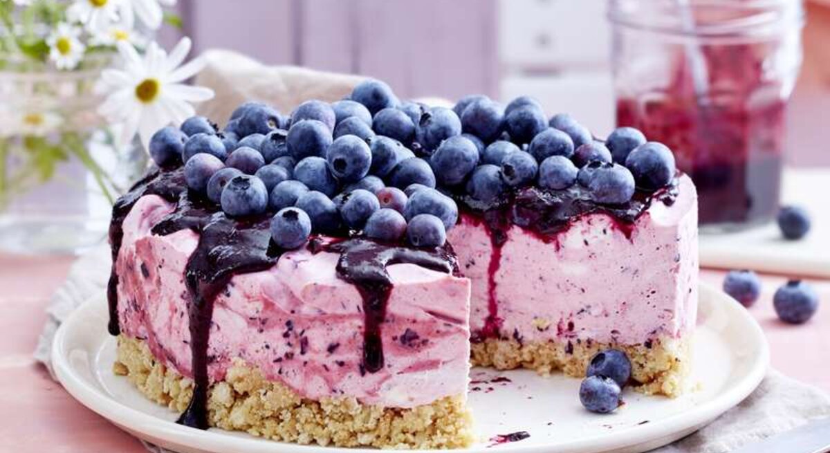 Heidelbeer-Cheesecake mit einem Topping aus frischen Heidelbeeren und einem Boden aus Butterkeksen, serviert auf einem hellen Tisch gedeckt mit rosa Tischdecke. 