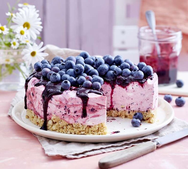 Heidelbeer-Cheesecake mit einem Topping aus frischen Heidelbeeren und einem Boden aus Butterkeksen, serviert auf einem hellen Tisch gedeckt mit rosa Tischdecke.  | © Cremiger Heidelbeer-Cheesecake für deine Gartenparty gefällig? 